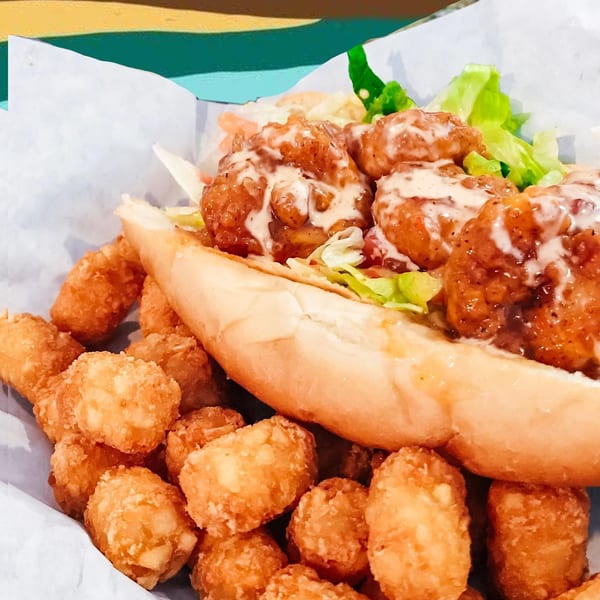 A shrimp po'boy sandwich with lettuce and sauce, served with a side of crispy tater tots.