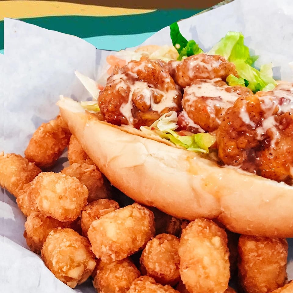 A shrimp po'boy sandwich with lettuce and sauce, served with a side of crispy tater tots.